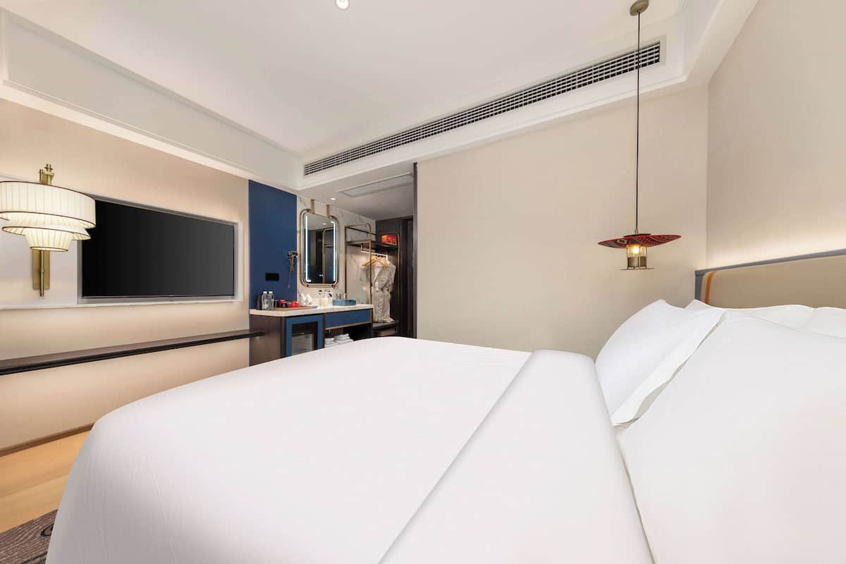 A modern hotel room with a large bed, a flat-screen TV mounted on the wall, a vanity with a mirror, and a hanging light fixture.