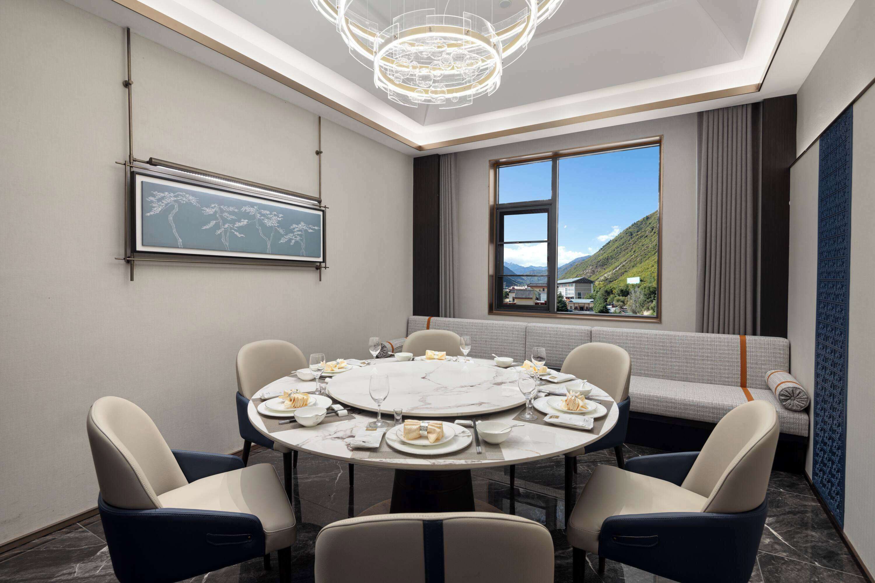 A dining room with a round table set for six, surrounded by upholstered chairs. A large window with a view of a mountain and a building is visible.