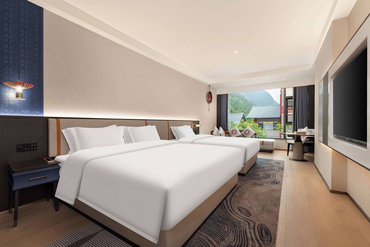 A modern hotel room with two beds, a desk, a TV, and a view of a mountain.