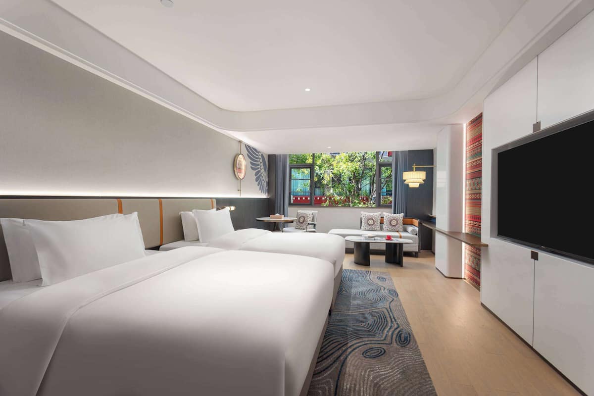 A hotel room with two beds, a TV, a seating area, and a view of greenery outside.