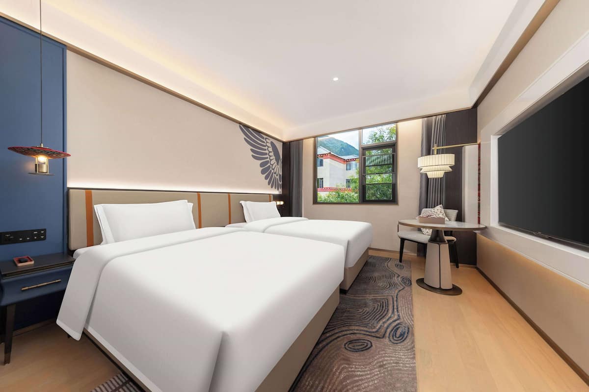 A modern hotel room with two beds, a desk, a chair, and a large window offering a view of a building and greenery.