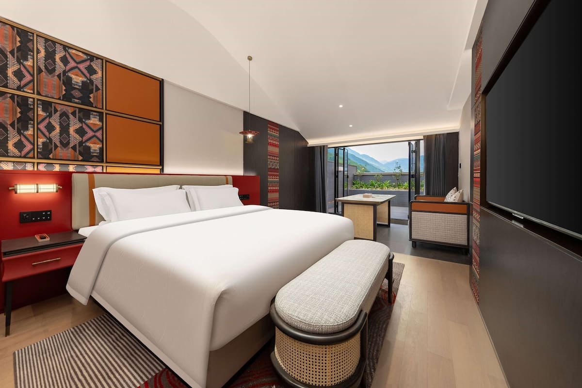 A modern hotel room with a large bed, a small seating area, and a view of mountains.