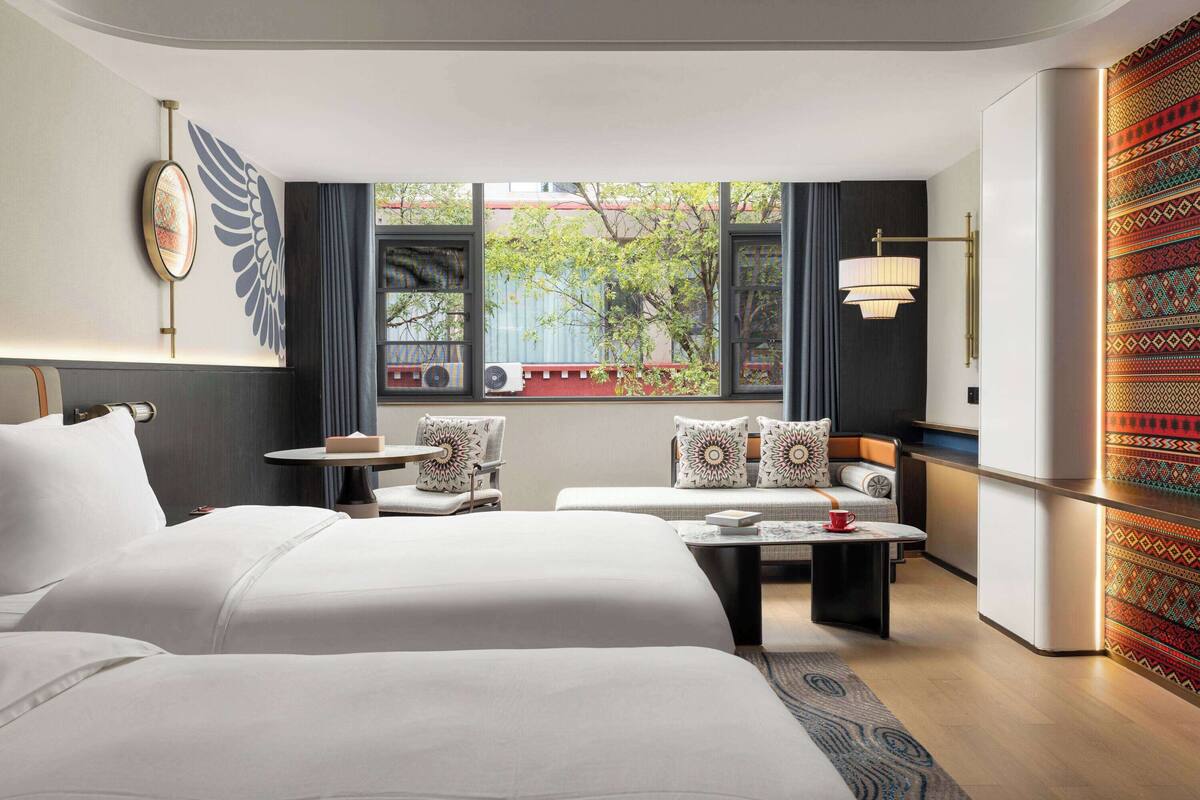 A hotel room with two beds, a small table, a sofa, and a view of the outside.