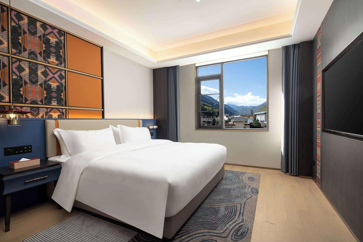 A modern hotel room with a large bed, a nightstand, a desk, and a large window offering a mountain view.