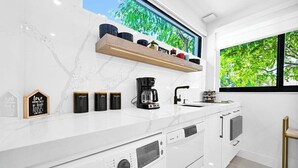 Una cocina moderna con cafetera, estante con frascos y ventana con vista a árboles.