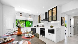 Una cocina moderna con armarios blancos, un protector contra salpicaduras de mármol y una zona de comedor con una mesa de madera dispuesta para cuatro comensales.