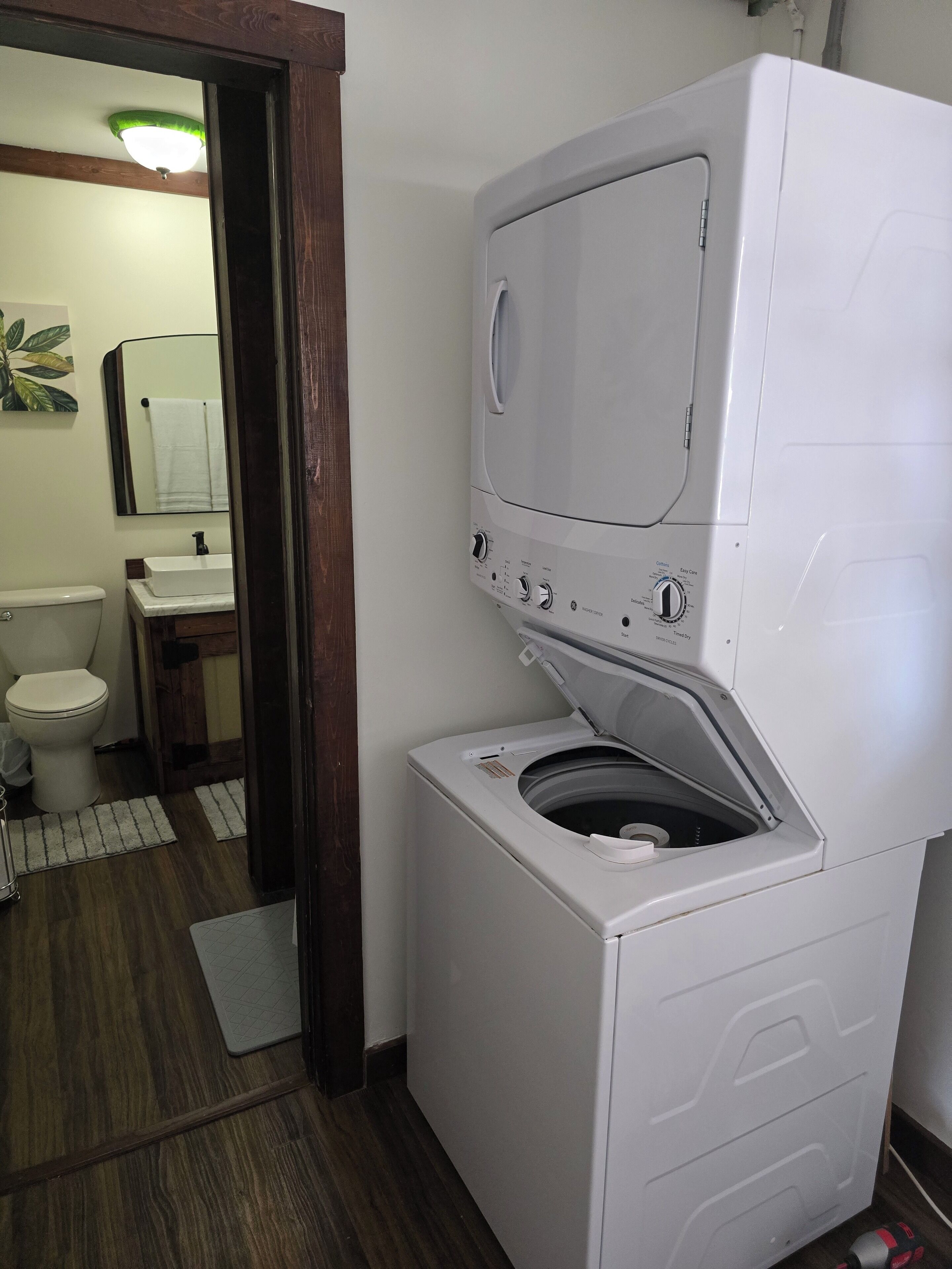 A compact laundry room with a front-loading washer and dryer, a wooden door, and a small sink with a mirror above it.