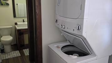A compact laundry room with a front-loading washer and dryer, a wooden door, and a small sink with a mirror above it.