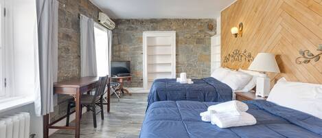 A hotel room with a stone wall, a wooden headboard, a desk with a chair, a television, and a bed with blue bedding and white towels.
