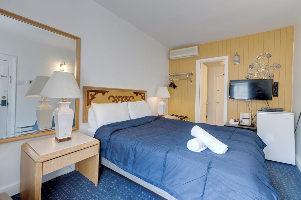 A hotel room with a bed, bedside lamps, a TV, a small refrigerator, and a mirror.