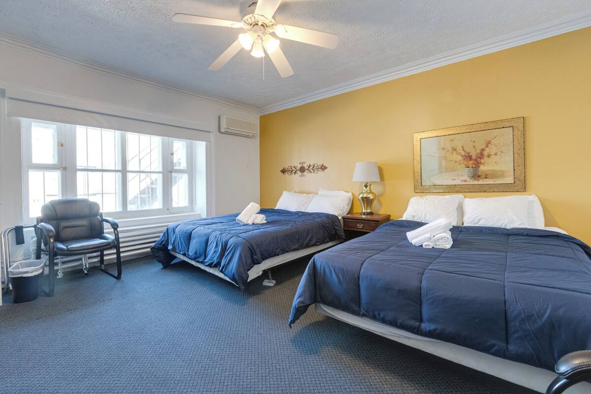 A hotel room with two beds, a chair, a lamp, a painting, and a window with a view.