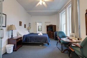 A hotel room with a bed, a desk, a chair, a TV, and a ceiling fan. - Hotel Le Cavalier - M11 (Québec)