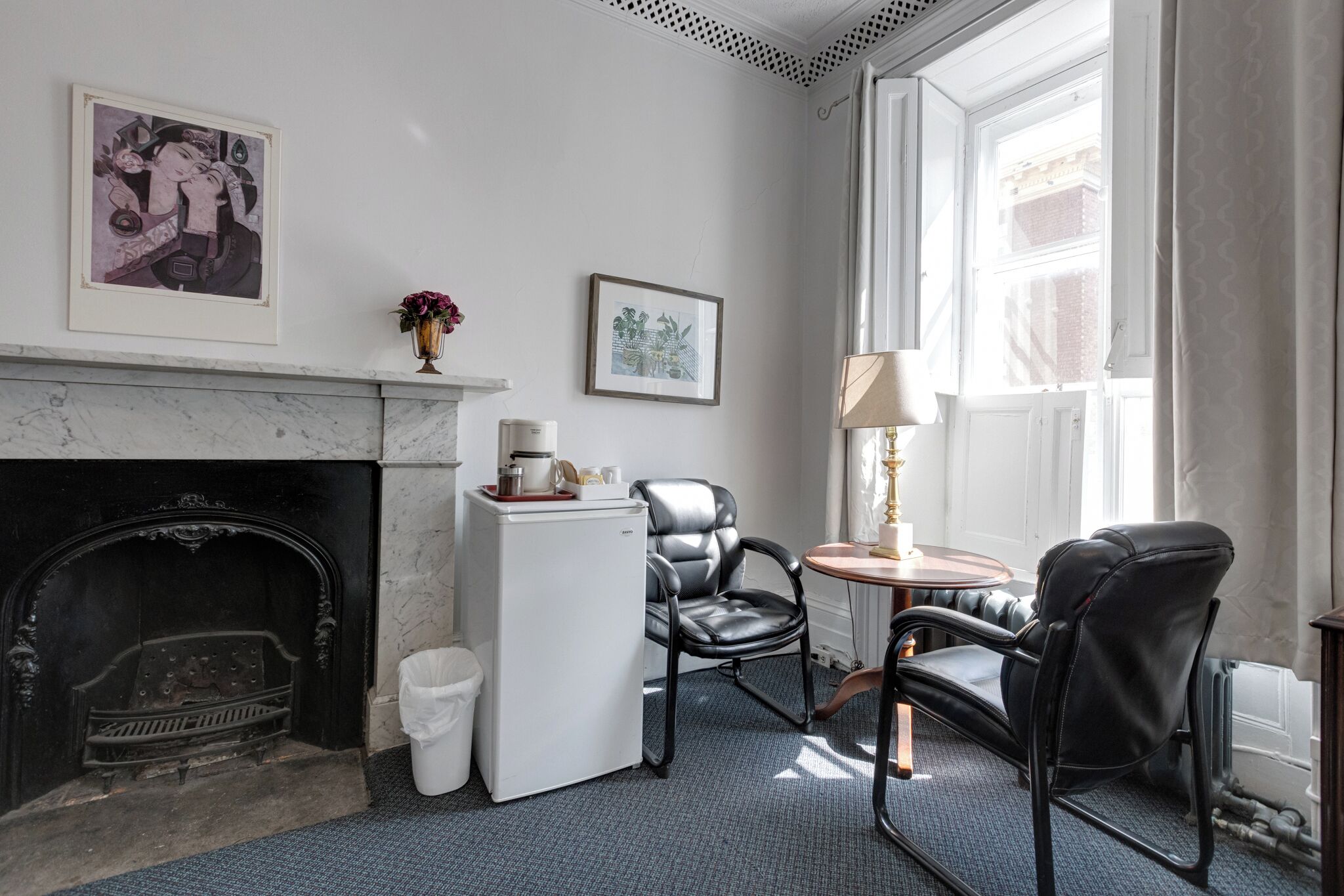 Une pièce avec une cheminée, un réfrigérateur blanc, deux fauteuils en cuir noir, une petite table avec une lampe et deux tableaux encadrés accrochés au mur.