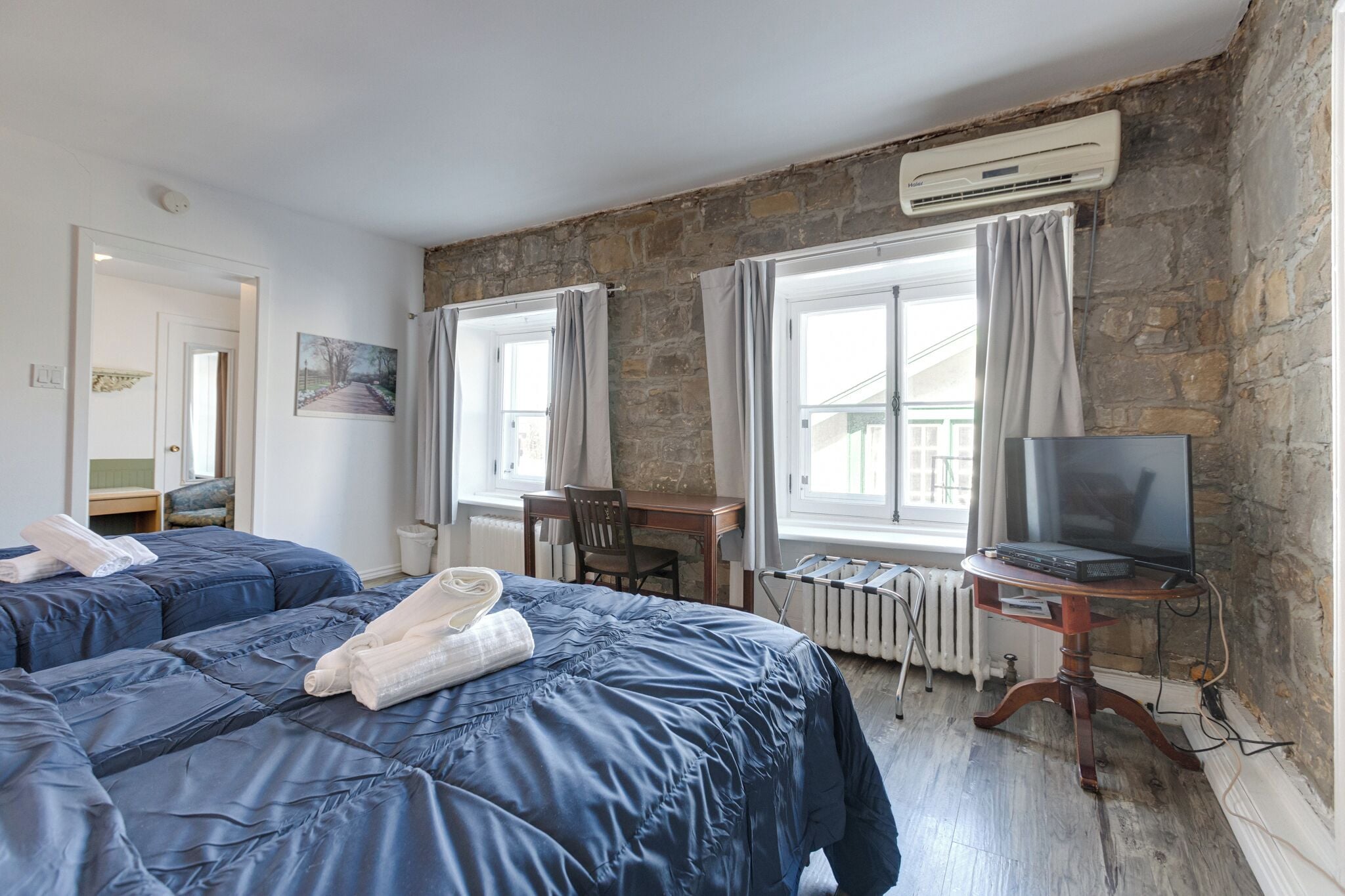 A hotel room with two beds, a desk, a chair, a television, and a stone wall.