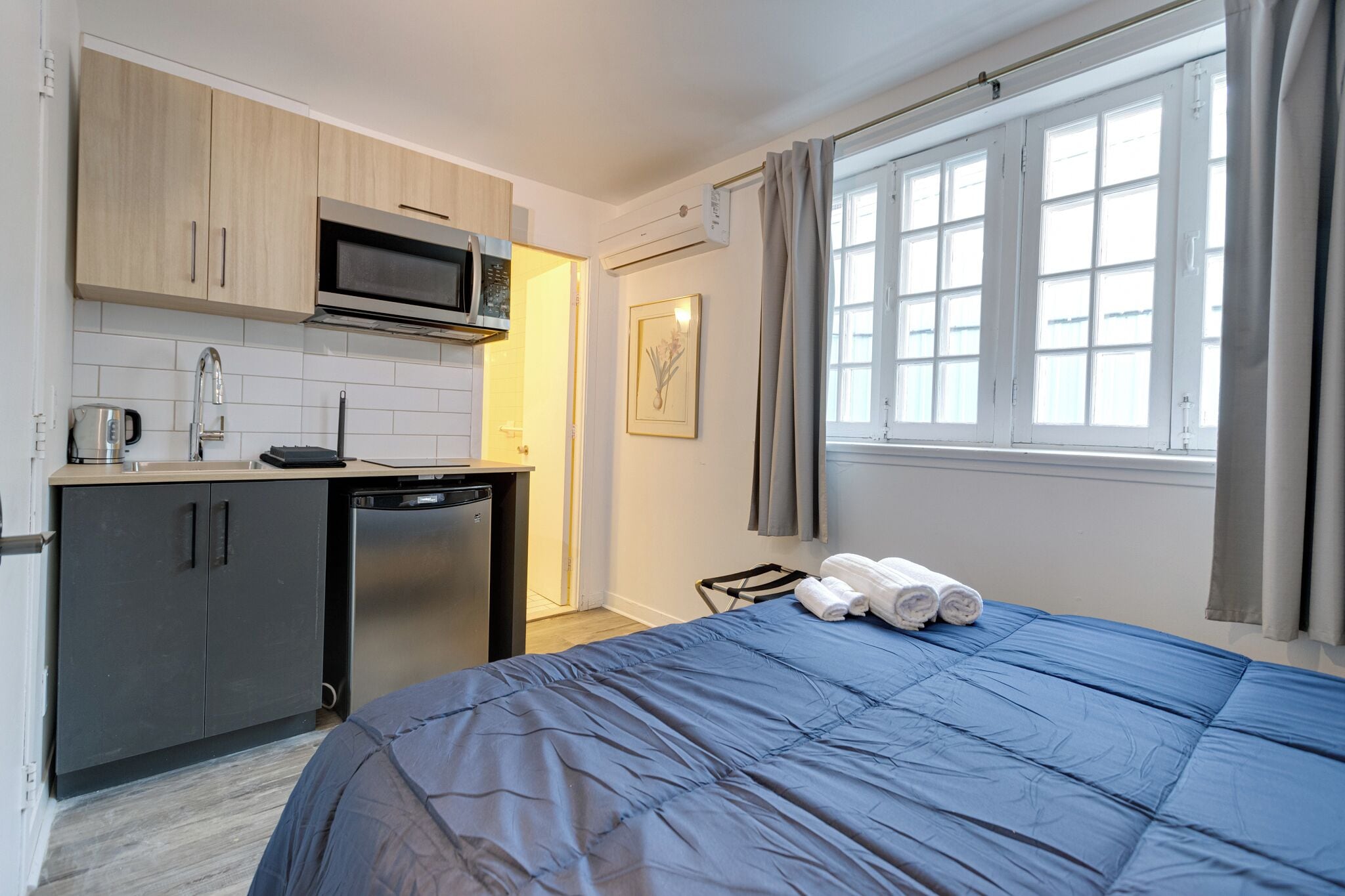 Una habitación de hotel compacta con kitchenette, una cama con cubrecama azul y una ventana con cortinas.