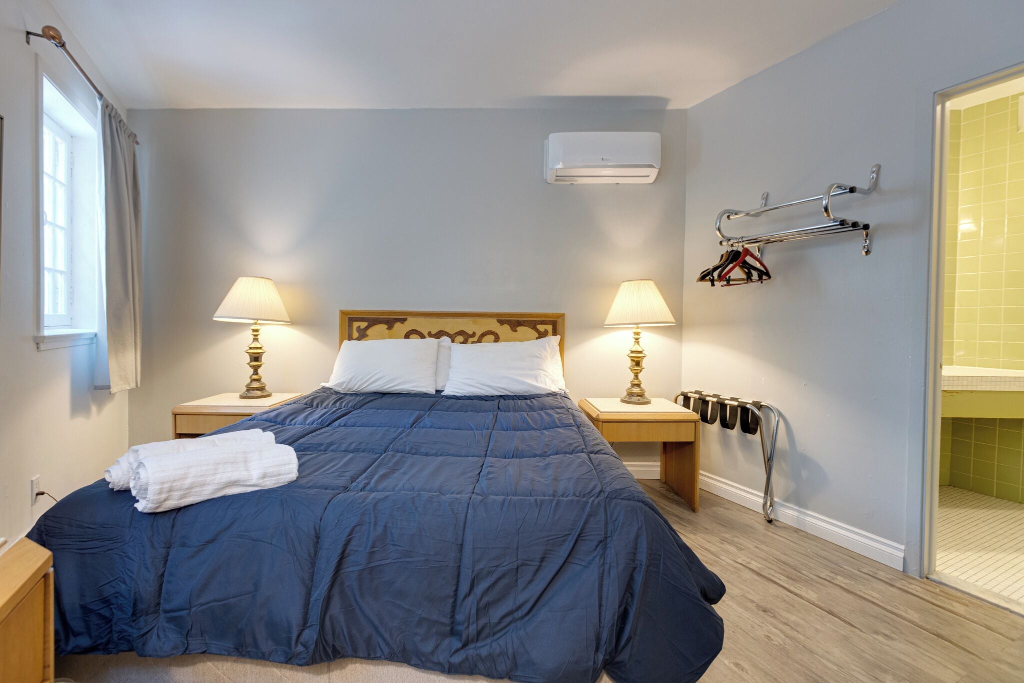 Een netjes opgemaakt bed met een blauwe deken en witte kussens, twee nachtlampjes, een wandairconditioner en een handdoek op het bed.
