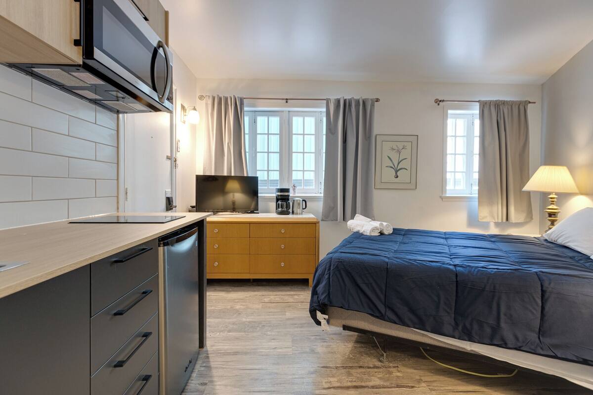 A compact bedroom with a bed, bedside table, lamp, and a view of the outdoors through a window.