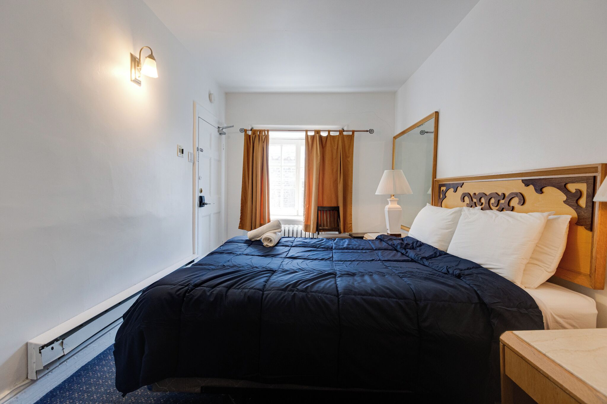 Una habitación de hotel con cama, mesita de noche, lámpara, ventana con cortinas y un espejo adosado a la pared.