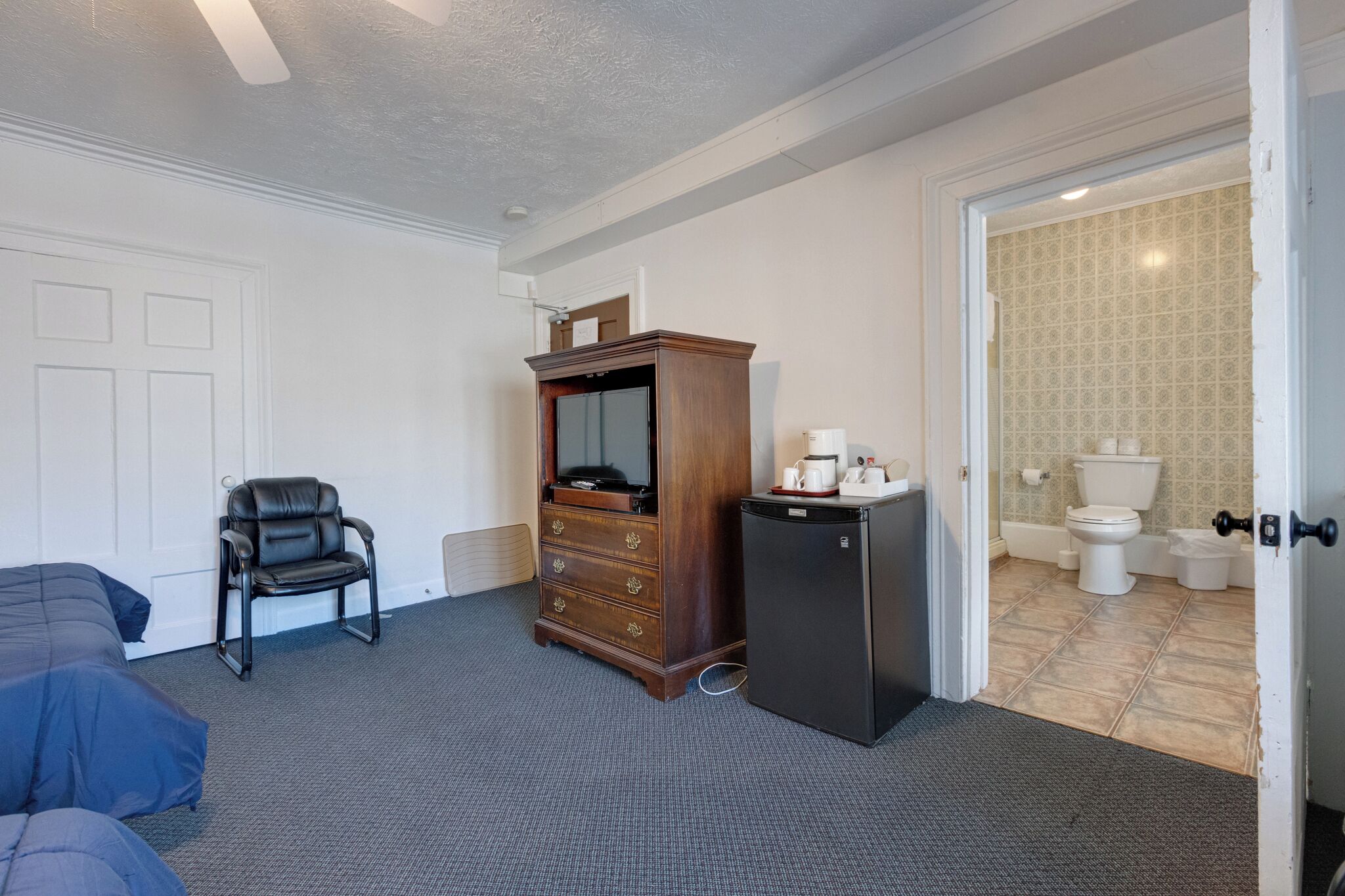 Ein Hotelzimmer mit einem Bett, einer hölzernen Kommode, einem schwarzen Kühlschrank, einem schwarzen Ledersessel und einem durch eine offene Tür einsehbaren Badezimmer.