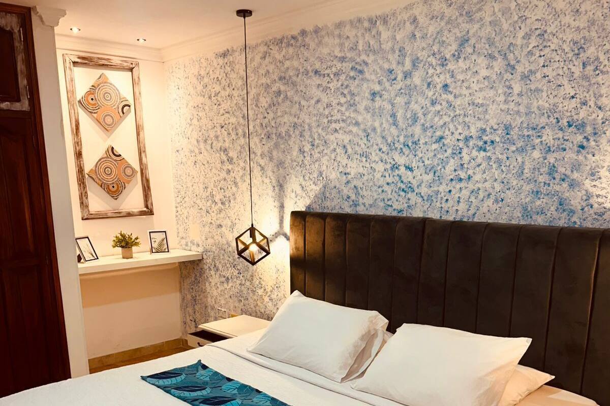 Una cama bien hecha con sábanas blancas y un plaid de hojas azules, un cabecero oscuro y una pared con un diseño texturizado en azul y blanco.