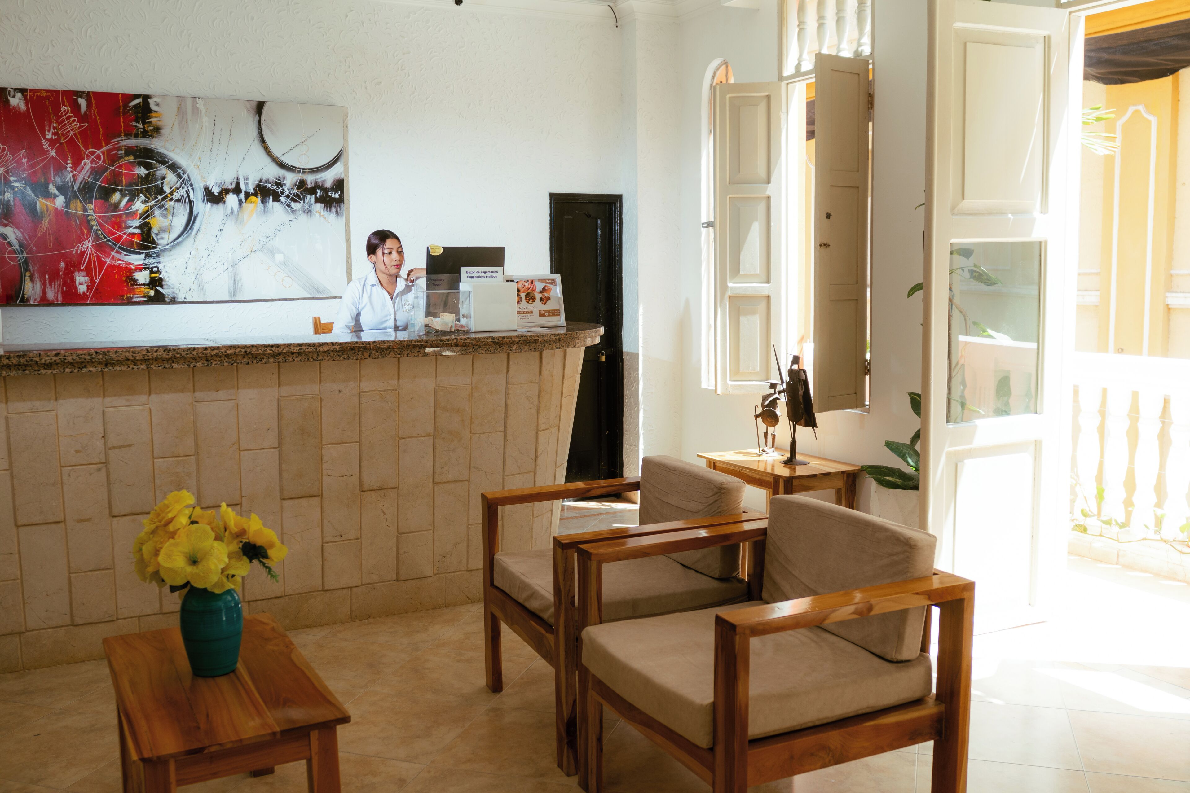 Un espace d’accueil comprenant un comptoir de réception, des chaises en bois, un vase avec des fleurs jaunes et un grand tableau abstrait accroché au mur.