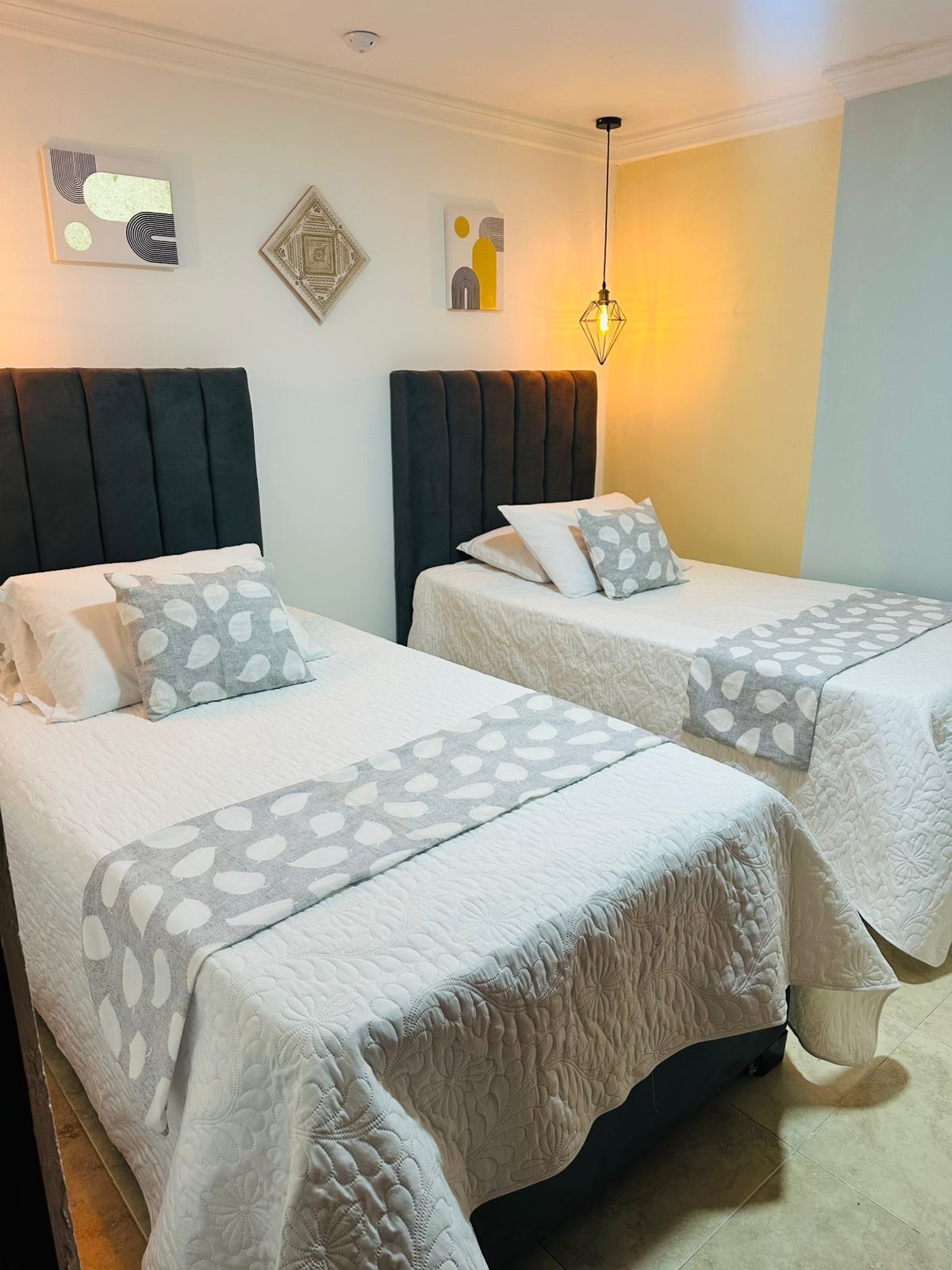 Una habitación de hotel con dos camas, cada una con cabecero y almohadas. Hay obras de arte en las paredes sobre las camas y una lámpara colgante arriba.