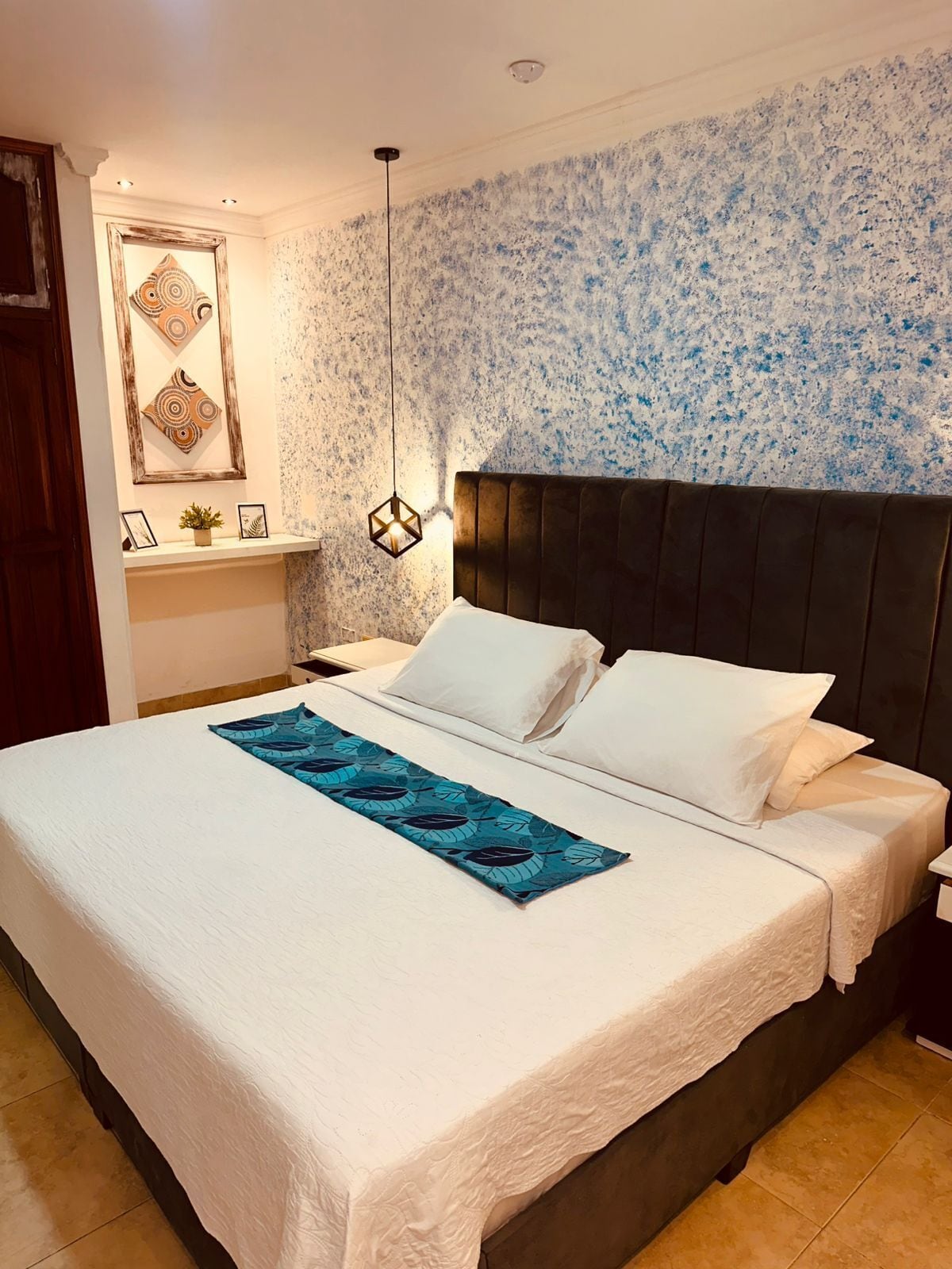 Un lit bien fait, avec du linge de lit blanc et un couvre-lit bleu orné de motifs de feuilles, une tête de lit sombre et un mur avec un motif texturé bleu et blanc.
