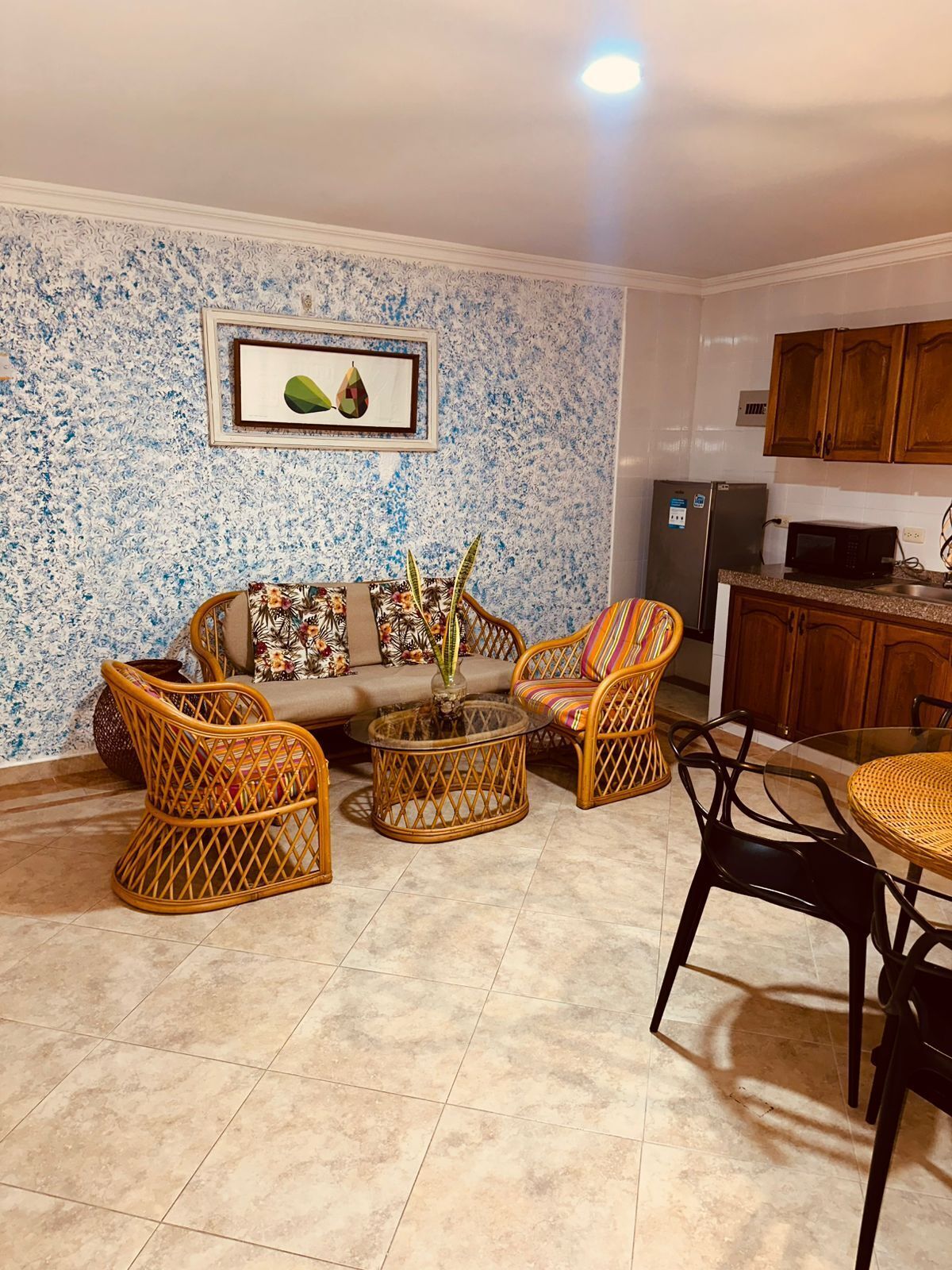 A living room with a sofa, two wicker chairs, a coffee table, and a dining table with chairs. There is a kitchen area with wooden cabinets and a refrigerator.
