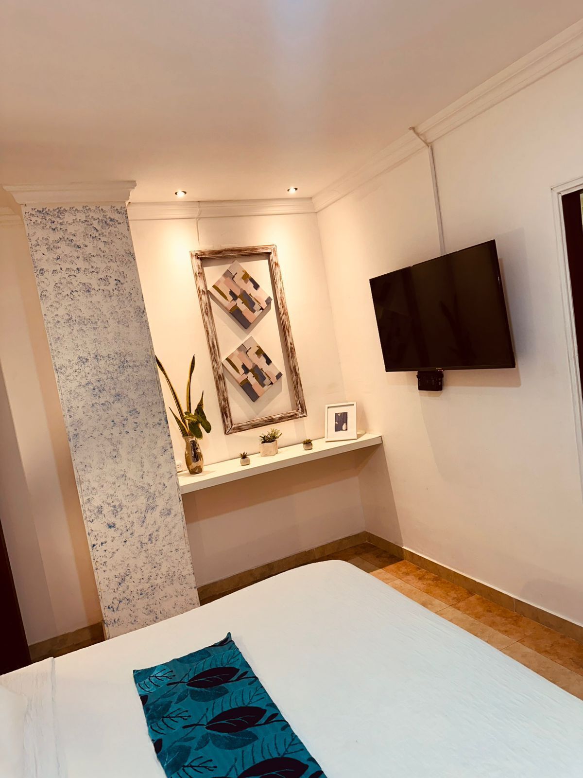 A hotel room with a bed, a TV mounted on the wall, a small shelf with decorative items, and a window with a floral curtain.