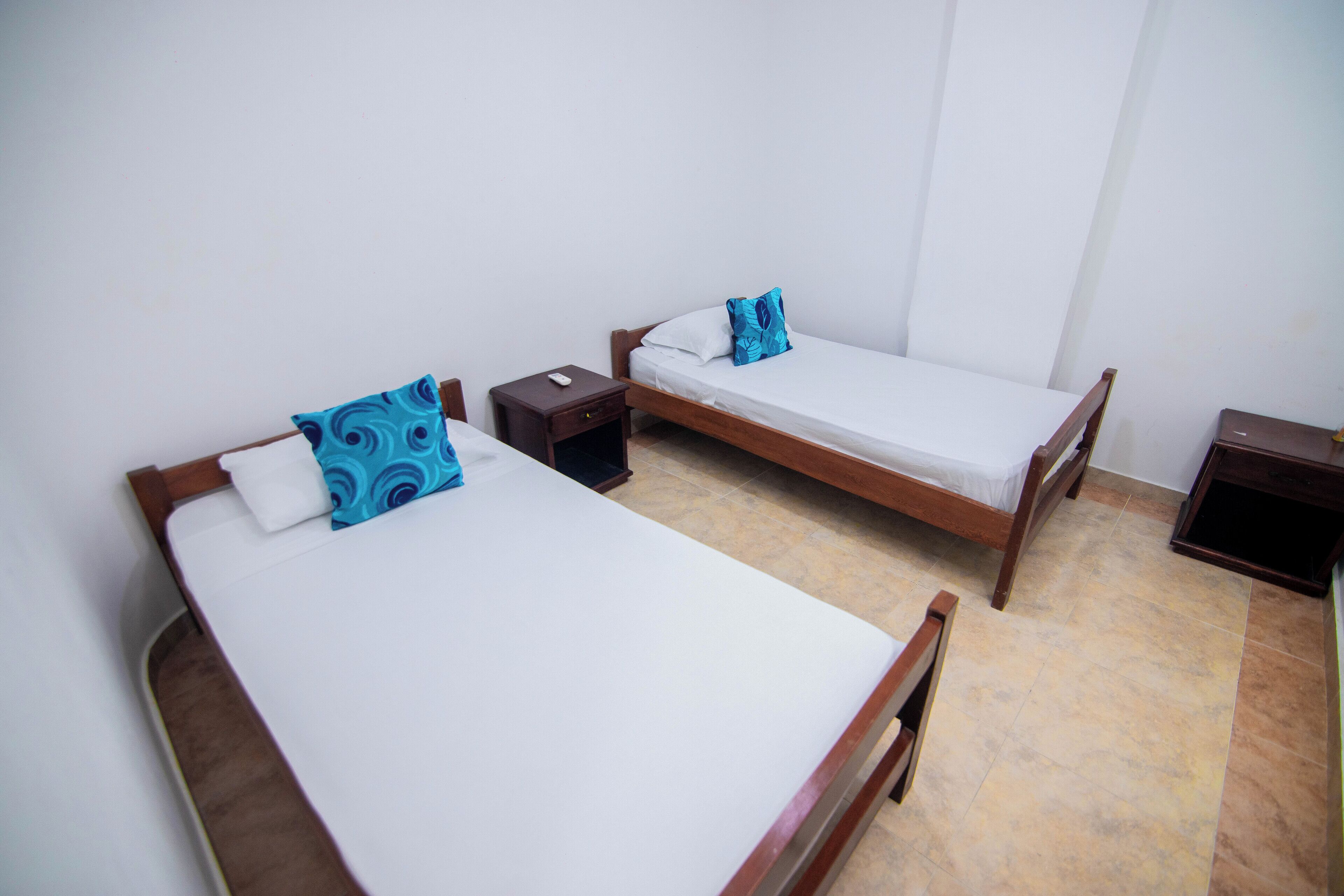 Duas camas de solteiro com estrutura de madeira e roupa de cama branca, cada uma com uma mesinha de cabeceira e um controle remoto.