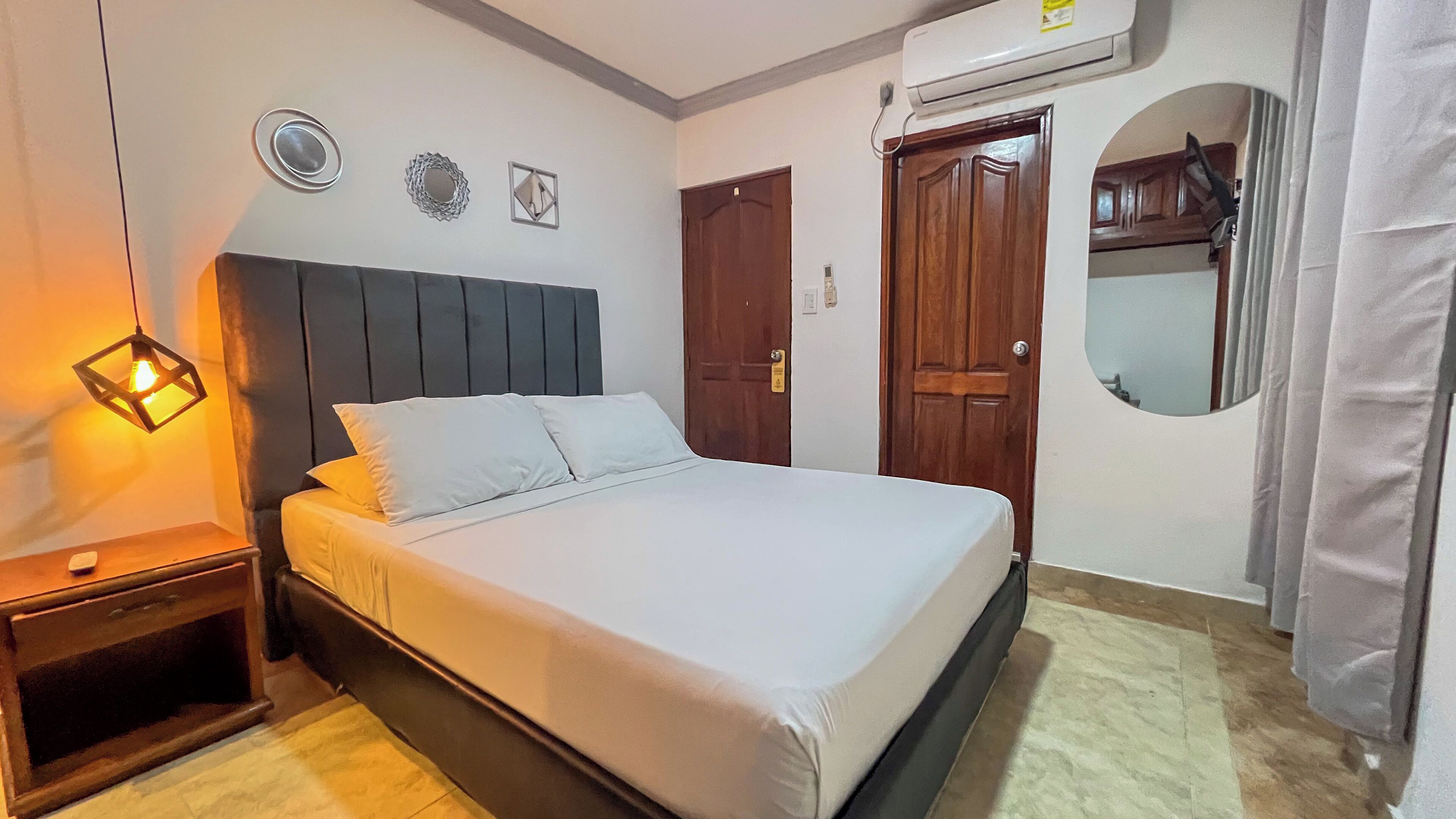 A neatly made bed in a room with a wooden headboard, a bedside table with a lamp, and a wall-mounted air conditioner.