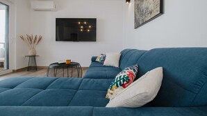A modern living room with a large blue sofa, a flat-screen TV mounted on the wall, a small round table with a vase of dried plants, and a large abstract painting. - Spacious Luxury Trogir Apartments - Standard Two Bedroom Apartment (Trogir)