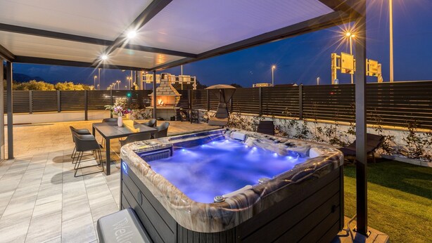 Un espacio al aire libre cubierto con un jacuzzi, asientos y vistas a la ciudad al anochecer.