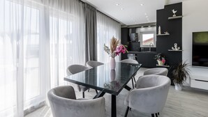 A modern dining area with a glass table and grey chairs, a kitchen in the background, and a large window with sheer curtains.