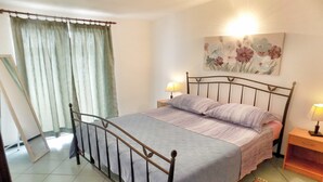 A bedroom with a metal bed frame, a single bed with a grey and purple bedspread, two bedside tables with lamps, a large floral painting on the wall, and a view of a balcony with curtains. - Trogir Sunny Apartments with Sea View - Standard Two bedroom Apartment with Sea view and Balcony (Trogir)