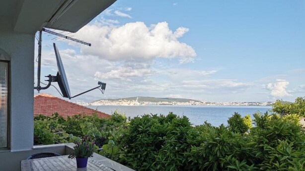 Un balcon avec une table et des chaises, un pot de fleurs et une vue sur la mer et les bâtiments éloignés.