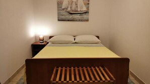 Une chambre à coucher comprenant un lit, une table de chevet, une lampe de chevet, un banc et un tableau représentant des voiliers accroché au mur.