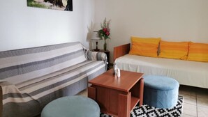 Un salon avec un canapé à rayures, une table basse en bois et un pouf bleu. On y trouve également un canapé blanc avec des coussins orange et un tapis à motifs noir et blanc. Un tableau représentant des arbres est accroché au mur, et un vase rempli de fleurs est posé sur une table d’appoint.
