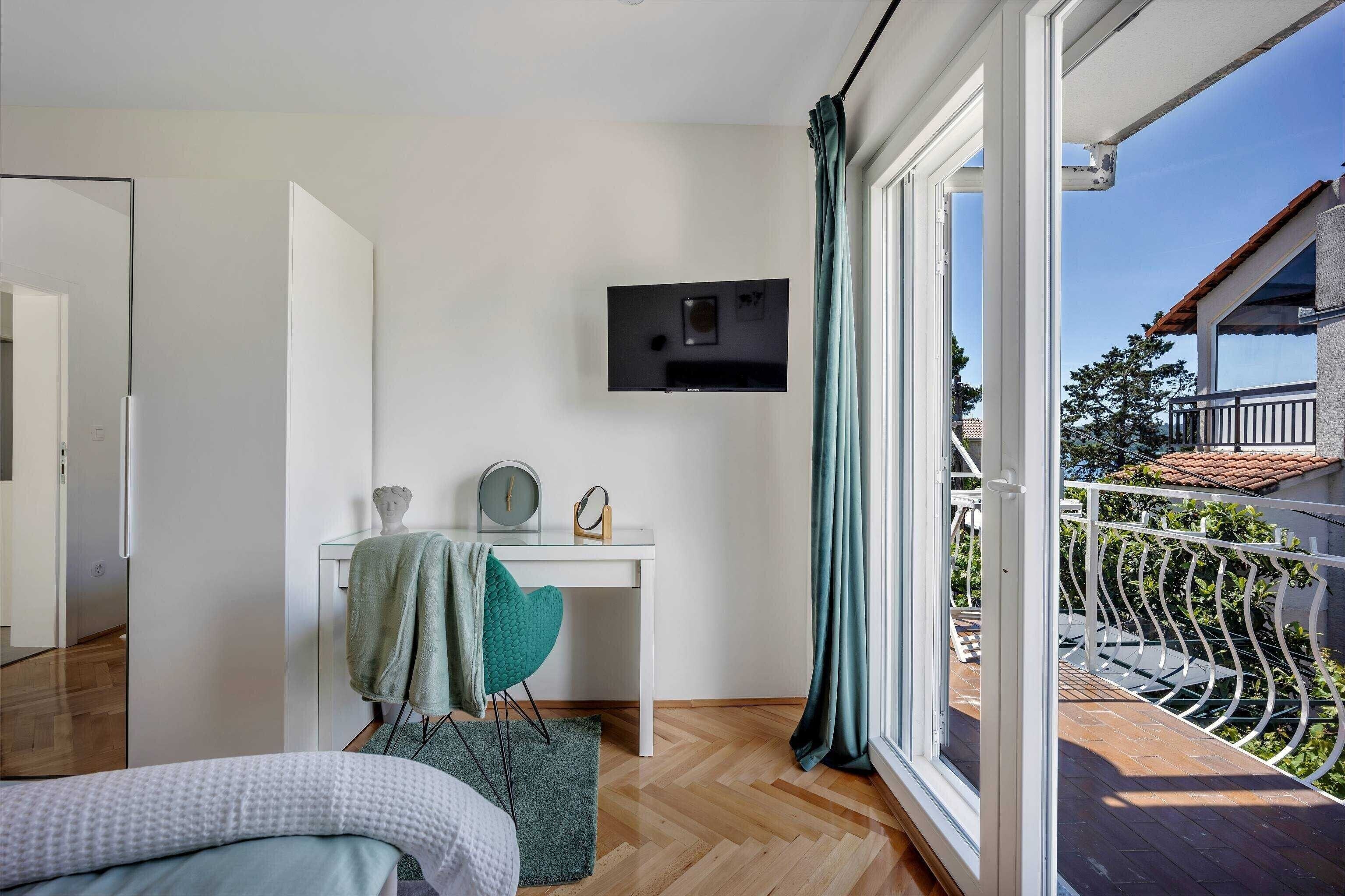Een slaapkamer met een bed, een bureau met stoel, een aan de muur bevestigde televisie en een balkon met een hek.