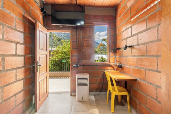 A small room with brick walls, a wooden door, a window with a view of greenery, a table with a chair, and a wall-mounted TV. - HOTEL EMBRUJO CAMPESTRE  (Jardin)