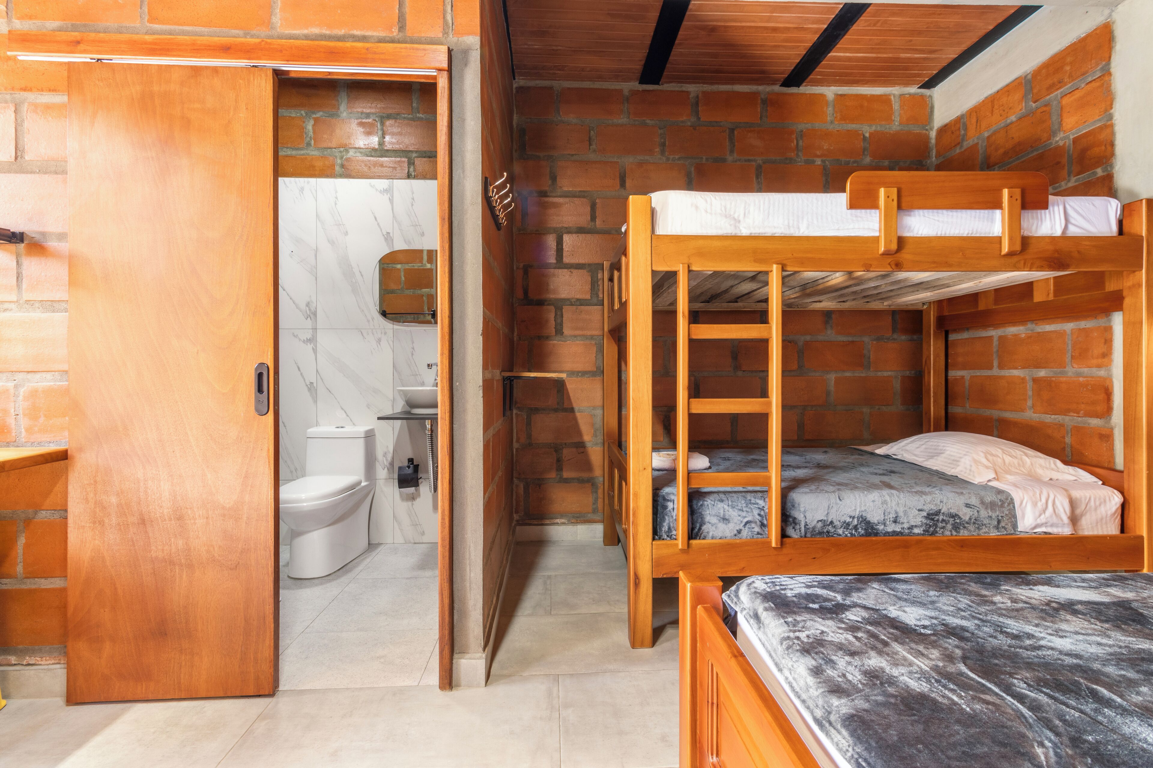Habitación pequeña con una litera de madera, un baño con inodoro y lavamanos, y una puerta que da a otra zona.