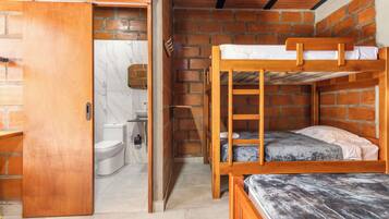 Habitación pequeña con una litera de madera, un baño con inodoro y lavamanos, y una puerta que da a otra zona.