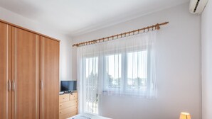 Un dormitorio con cama, un armario de madera, un televisor sobre una cómoda de madera y una ventana con cortinas traslúcidas.
