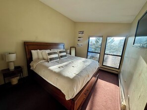 A bedroom with a large bed, a nightstand, a lamp, and a window with a view of trees. - Comfortable Mountain Condo Close to Everything! (Dillon)