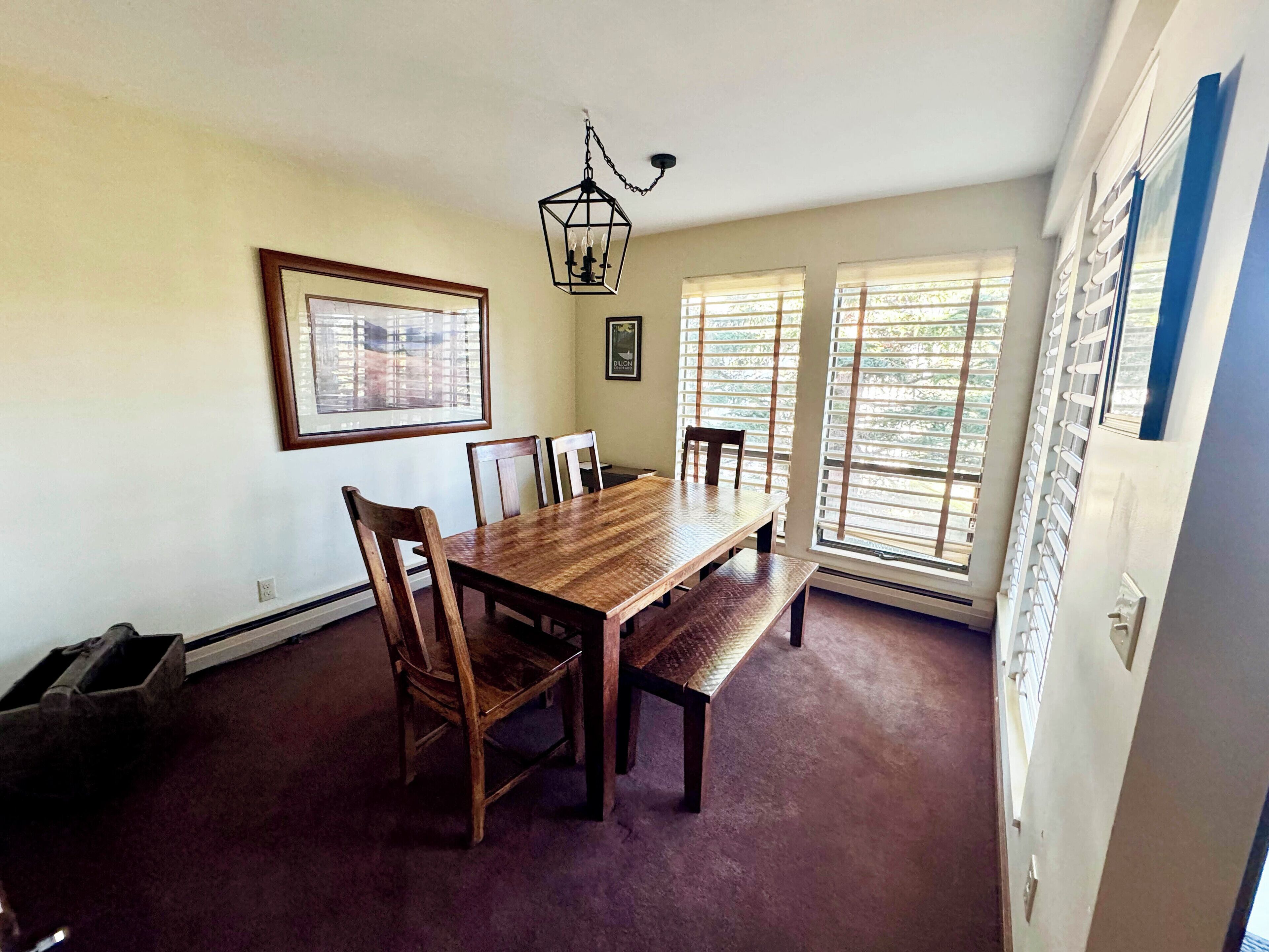 A dining room with a wooden table and chairs, a chandelier, and large windows with blinds.