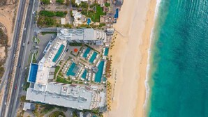 Eine Vogelperspektive eines Küstenhotels mit mehreren Schwimmbecken, einem Strand und einer parallel zur Küste verlaufenden Straße.