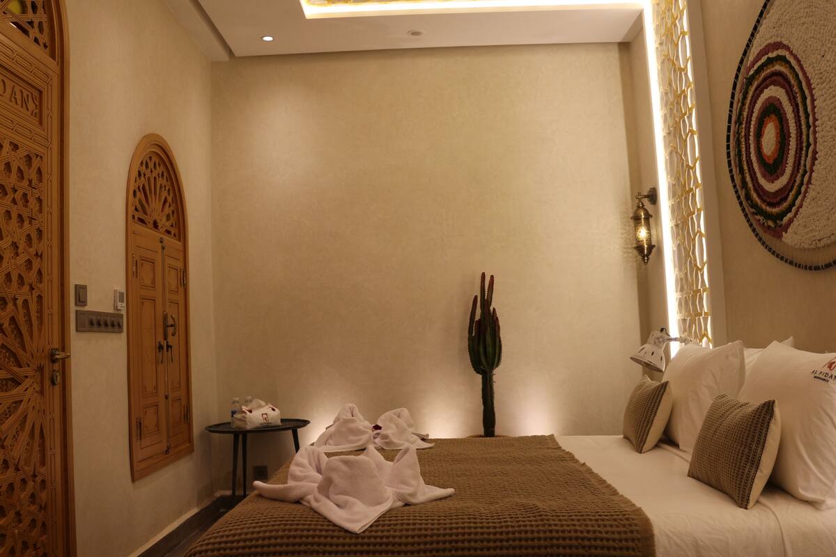 A hotel room with a bed, a small table with towels, a cactus, and a decorative wall hanging.