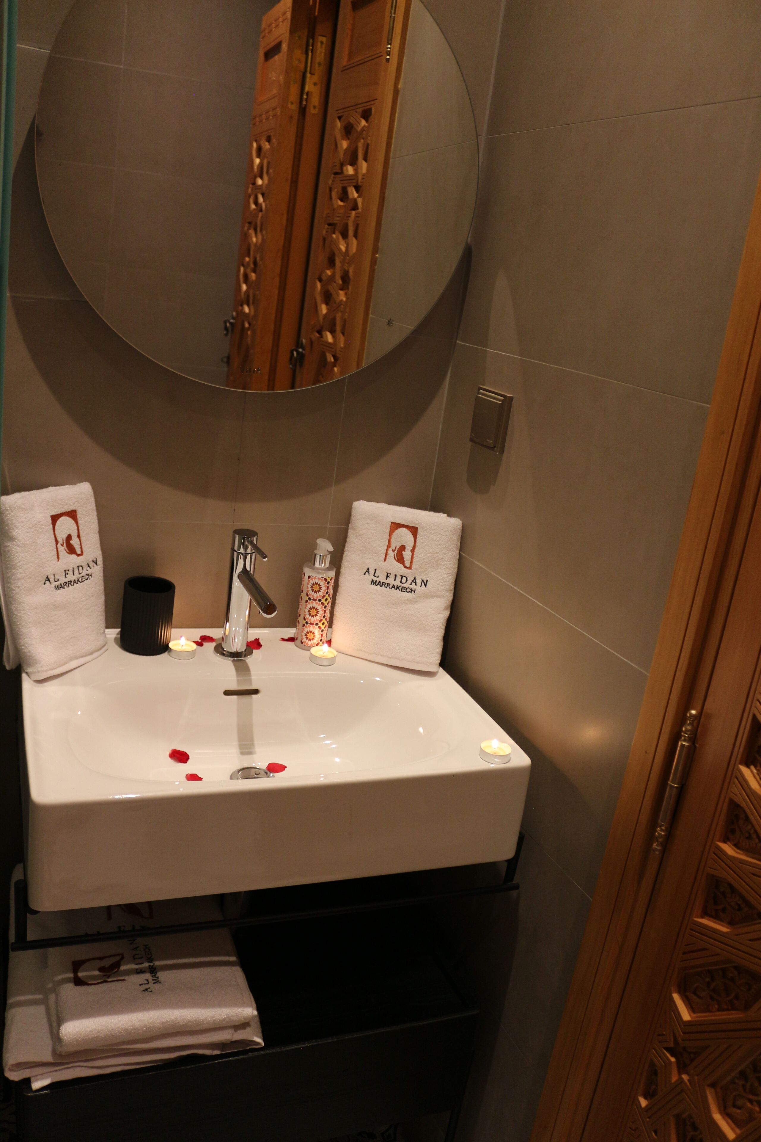 Une salle de bain avec un lavabo blanc, deux serviettes ornées du logo « Al Fidan Marrakech », un miroir rond et une porte en bois.