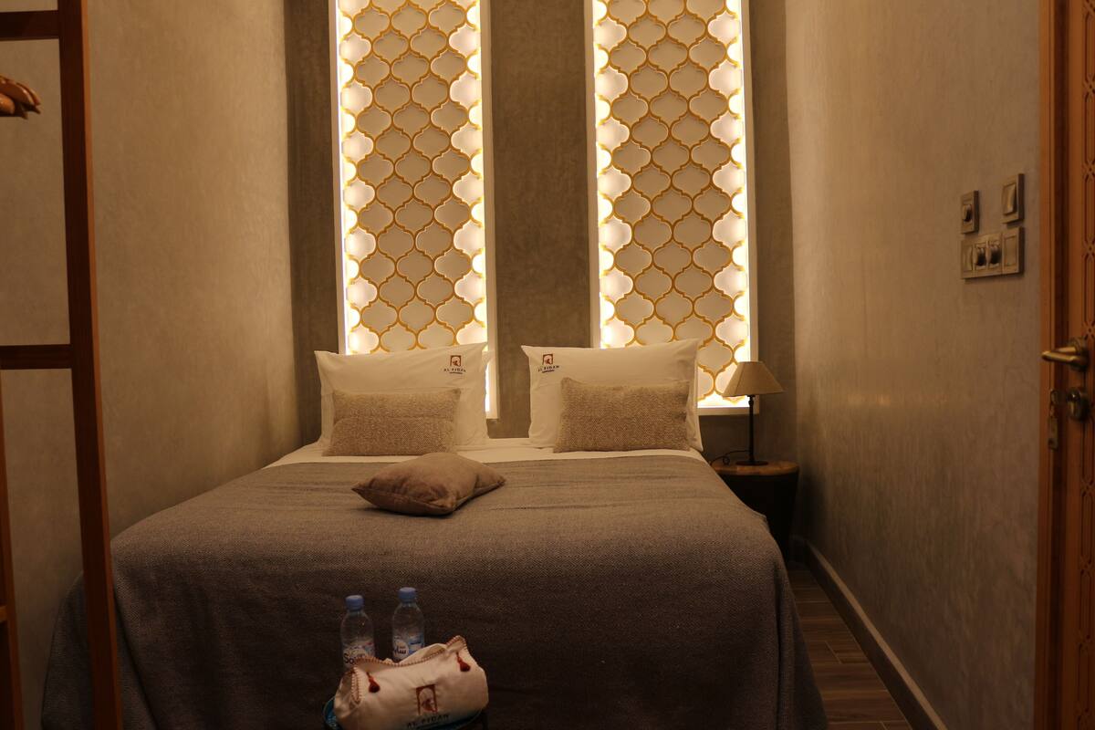 A hotel room with a bed, pillows, a nightstand with a lamp, and a water bottle on the floor.