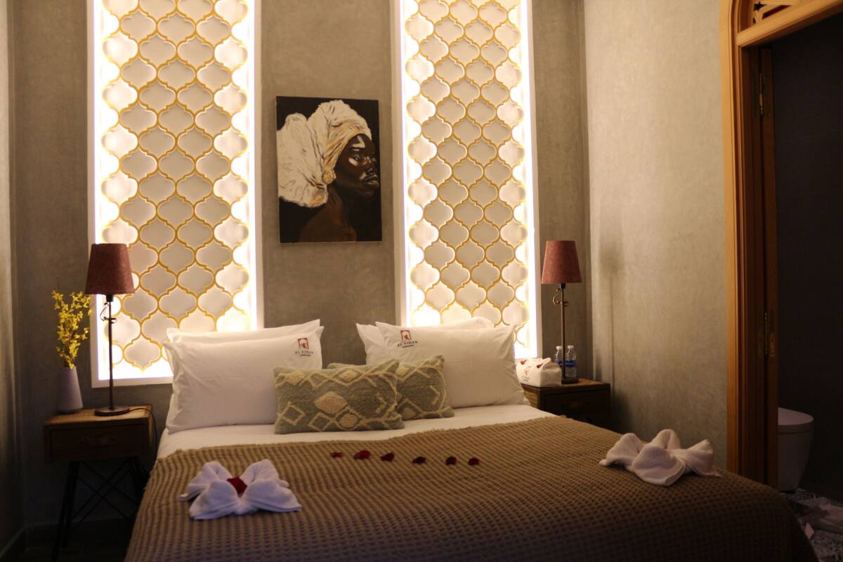 A hotel room with a bed, pillows, a painting, and a window with a patterned curtain.